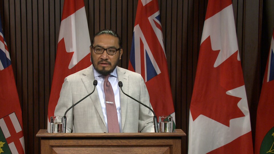 Un homme parle à la tribute devant des drapeaux canadien et ontarien.