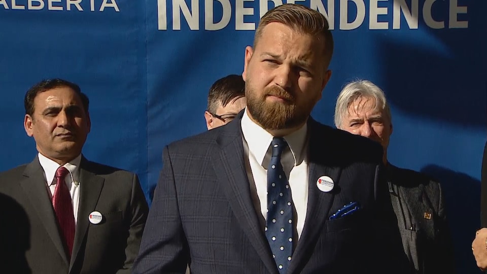 Un homme avec une barbe. Il porte un costard avec une cravate bleue à pois blancs. Trois autres hommes sont situés derrière lui. On aperçoit au fond une banderole qui indique en anglais: « Alberta » et « independence ».