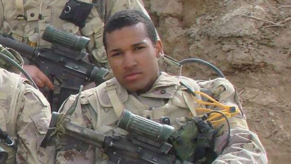 Lionel Desmond en uniforme militaire assis en Afghanistan, son fusil d'assaut repose sur ses genoux.