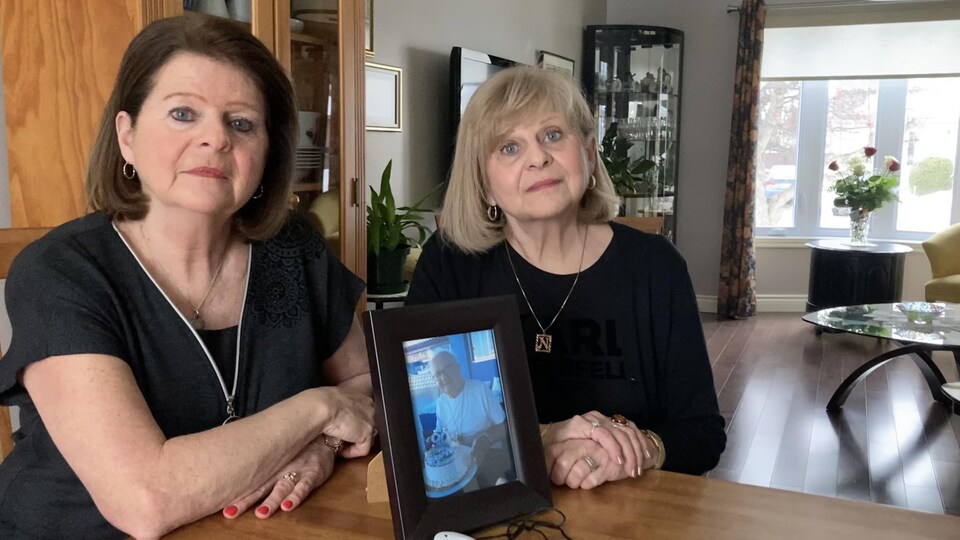 Louise et Ninon Comtois sont assises à une table avec, devant elle, une photo de leur père et l'appareil de détection de chute qu'il avait l'habitude de porter. 