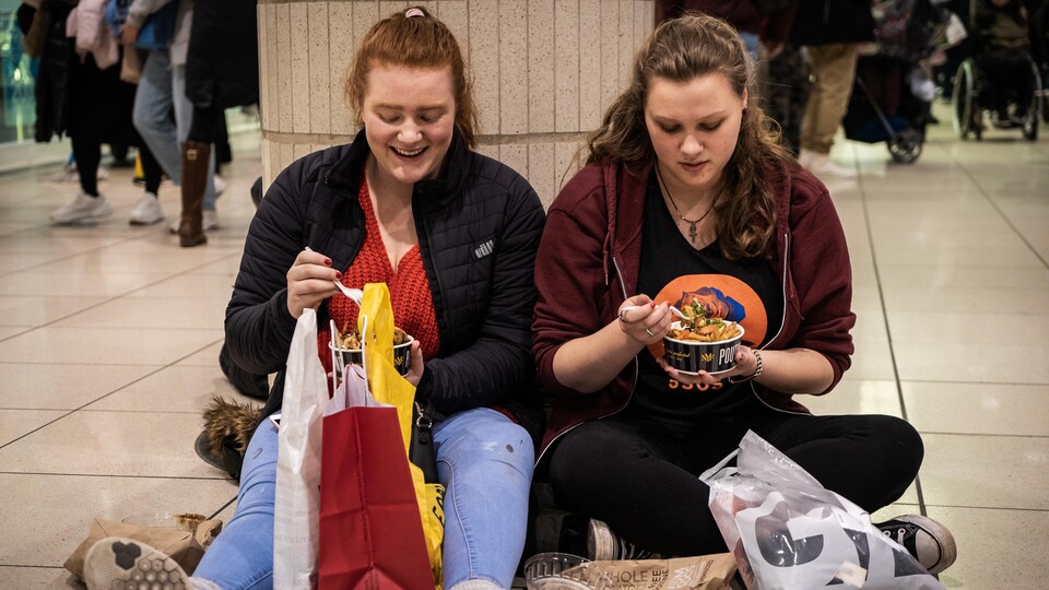 Deux jeunes filles dÃ©gustent une poutine assises au sol au milieu d'une galerie marchande.