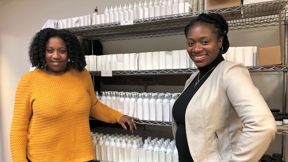 Deux femmes d'origine haïtienne, l'une en pull jaune, l'autre en veste blanche et pull noir, devant une étagère de flacons 