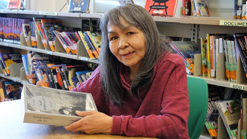 Une femme aux cheveux longs gris tient un livre dans ses mains, assise à une table dans une bibliothèque