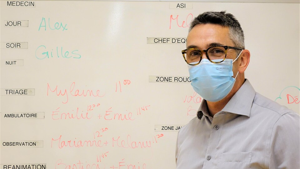 Un monsieur à lunettes avec masque de procédure devant un tableau blanc où sont inscrits les noms du personnel en poste