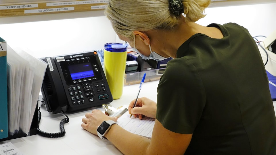Une jeune femme aux cheveux blonds attachés, de dos, en train d'écrire sur un calepin à côté d'un téléphone.
