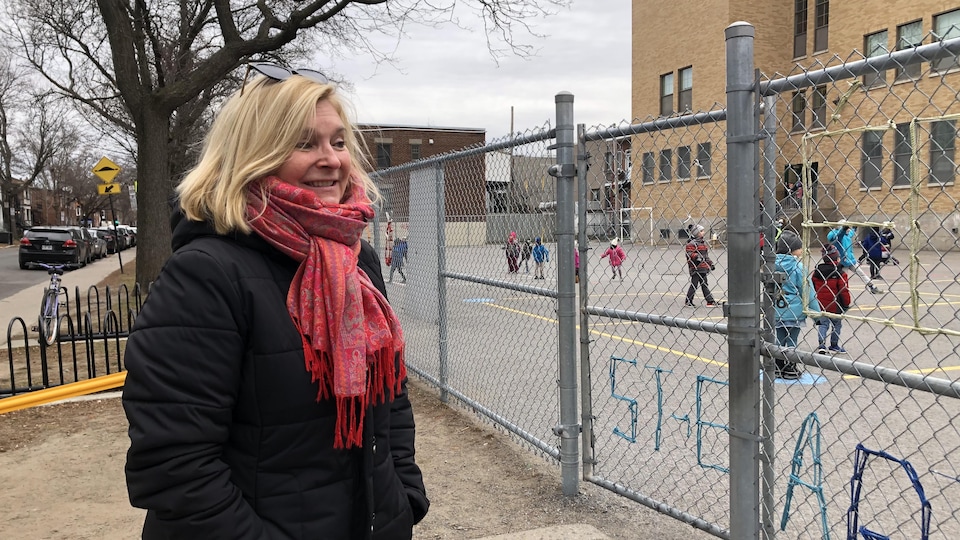 The teacher with blond hair, black coat, pink scarf, next to the fence of a schoolyard