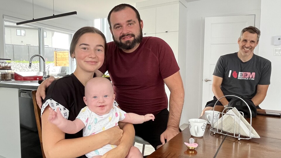 Un couple avec un bébé et un homme à droite qui sourit en regardant la petite famille.