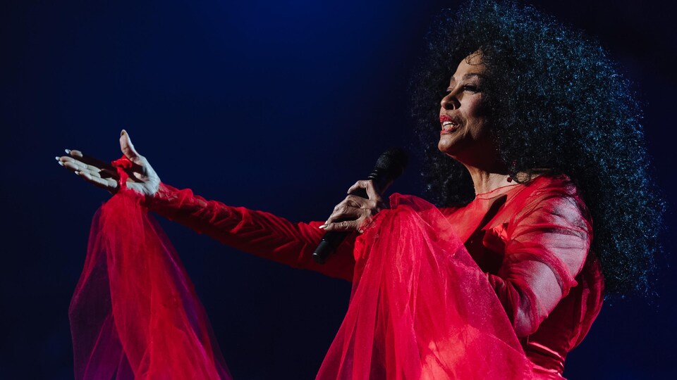 La chanteuse porte une robe rouge et lève le bras en chantant dans un micro sur scène.