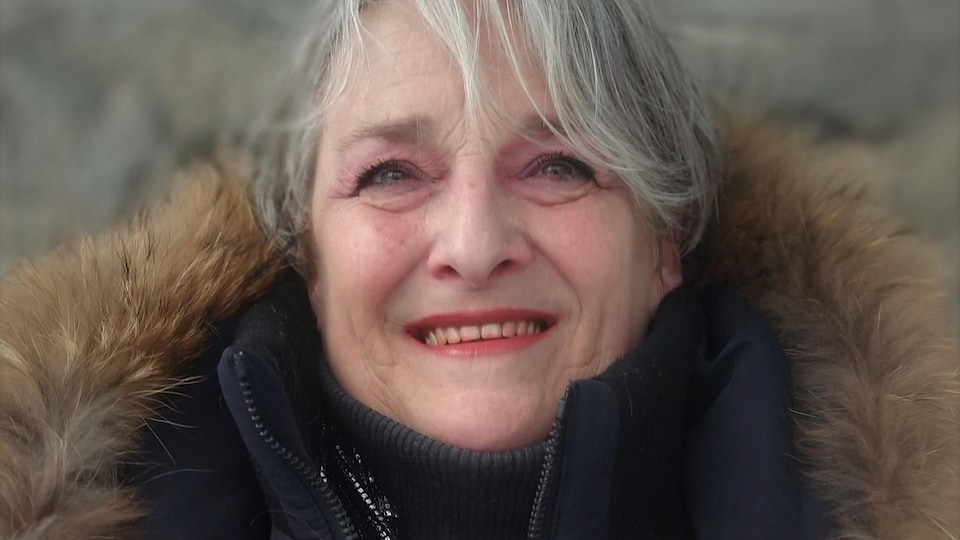Portrait en couleur, en extérieur, d'une femme aux cheveux gris portant un manteau noir avec une bordure de fourrure. Elle regarde au loin, face à l'appareil photo, et sourit.