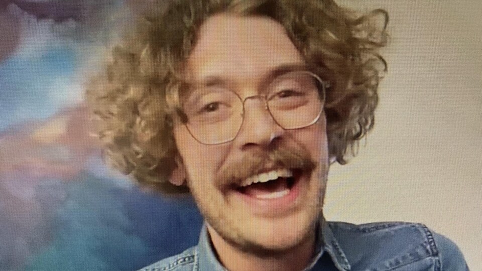 Capture d'écran : un jeune homme souriant aux cheveux bouclés portant des lunettes