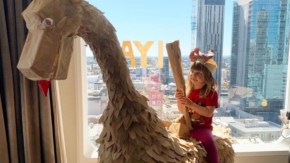 Une jeune enfant est assise sur un dinosaure en papier dans une chambre d'hôtel.
