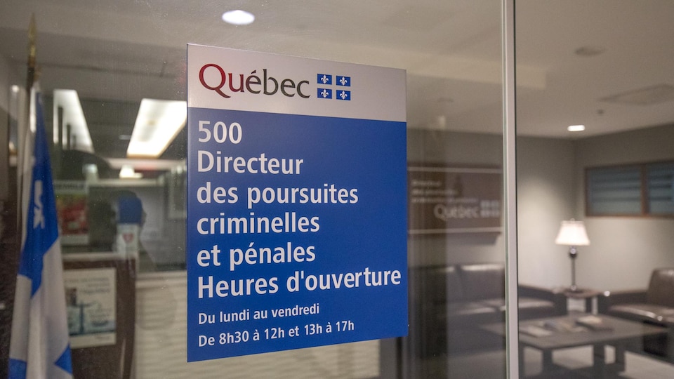 Une affiche du Directeur des poursuites criminelles et pénales est posée sur une cloison de verre. De l’autre côté de la cloison, on aperçoit un drapeau du Québec, un comptoir d’accueil, des fauteuils ainsi qu’un table basse.