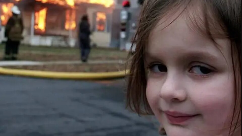 Une petite fille regarde l'objectif de la caméra avec un air coquin alors qu'un feu ravage une maison derrière elle.
