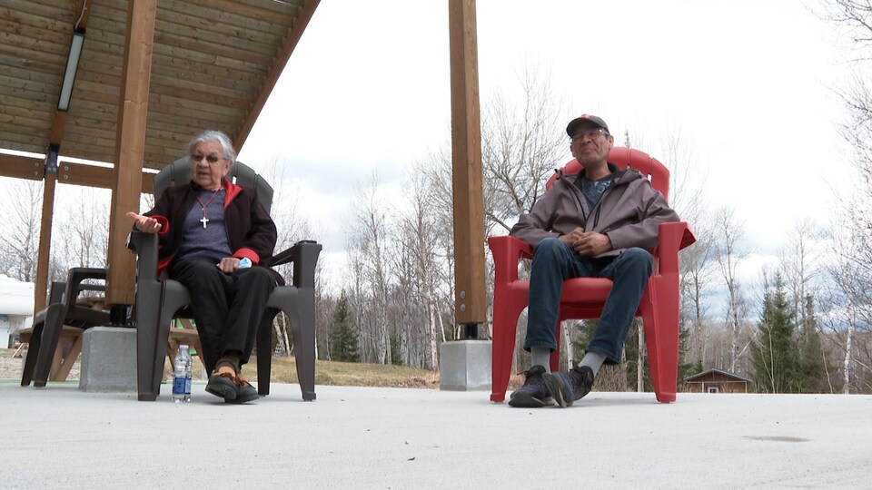 Deux personnes d'origine autochtone assises dans des fauteuils Adirondack près d'un chalet.