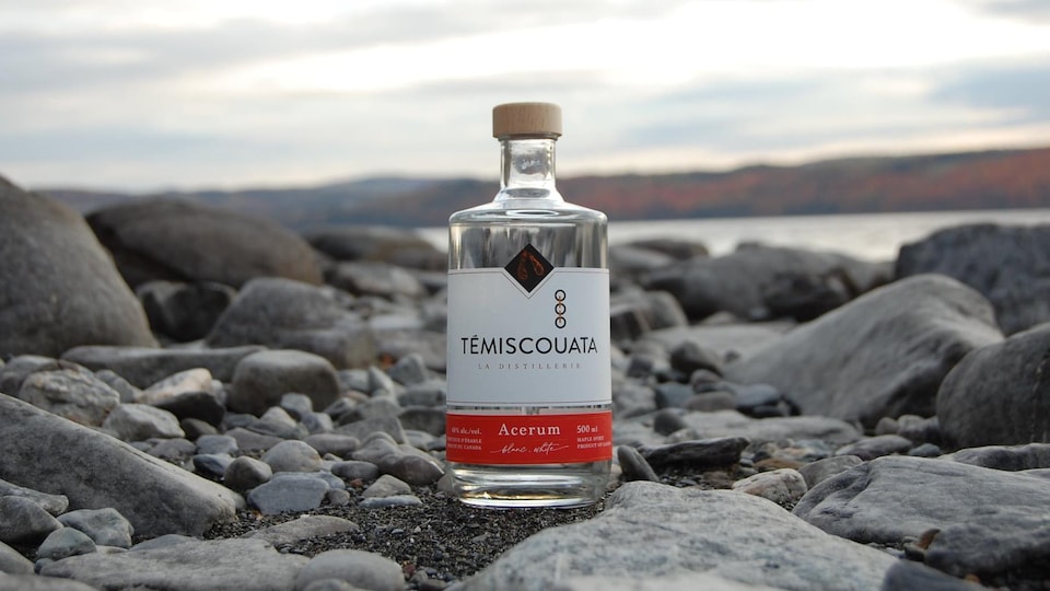 Une bouteille de l'acerum blanc de la Distillerie Témiscouata est déposée sur les roches, près d'un lac.