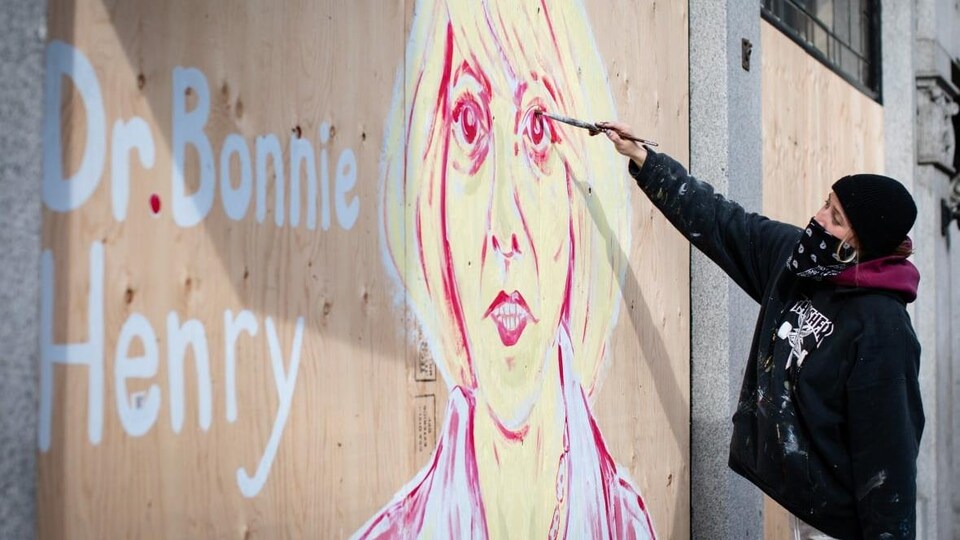 Une femme peint une image de Bonnie Henry sur une plaque de bois recouvrant une vitrine de boutique fermée dans le quartier Gastown.