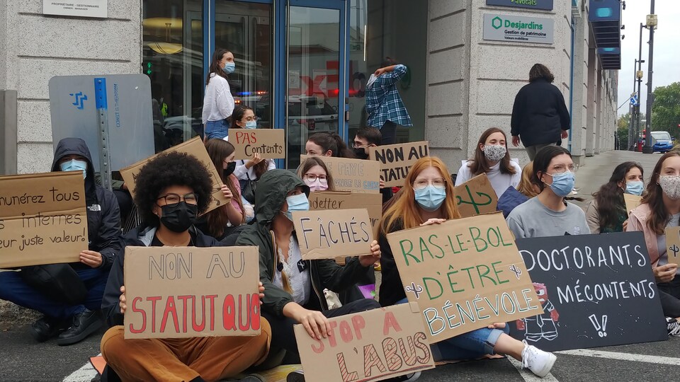 Des étudiants tenant des affiches sont assis sur le sol devant l'entrée de l'édifice Royale abritant le bureau du député de Trois-Rivières.