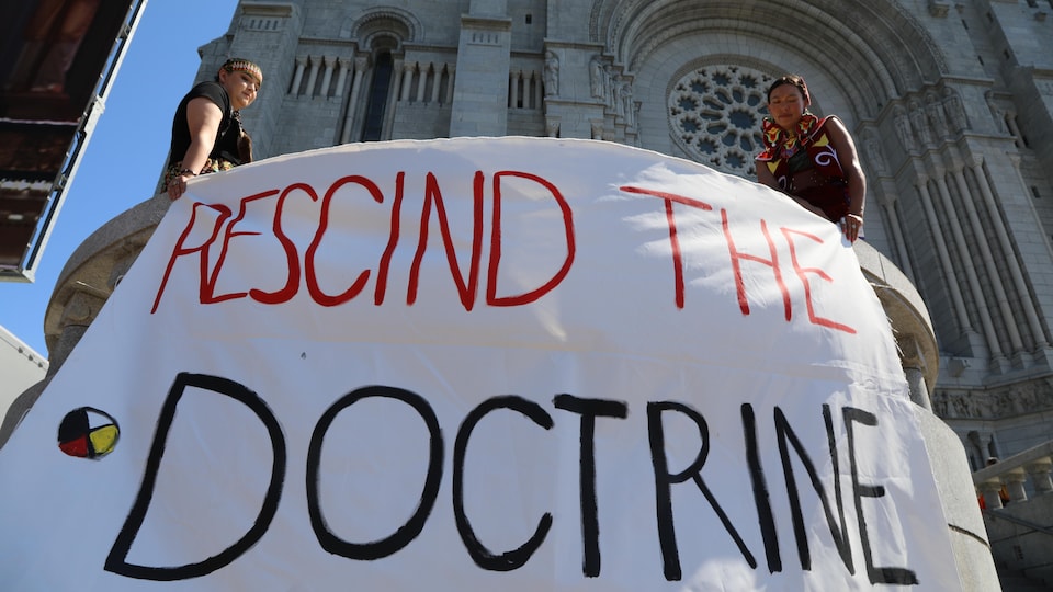 Deux personnes autochtones tiennent une banderole où il est écrit, en anglais « Abrogez la doctrine ».