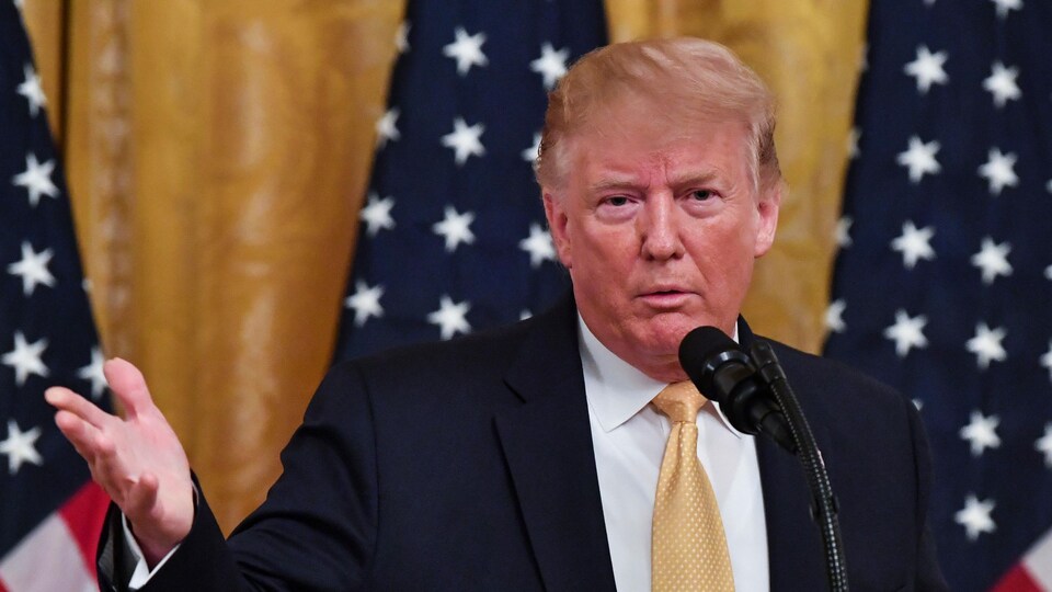 L'homme parle au micro avec trois drapeaux américains en arrière-plan.