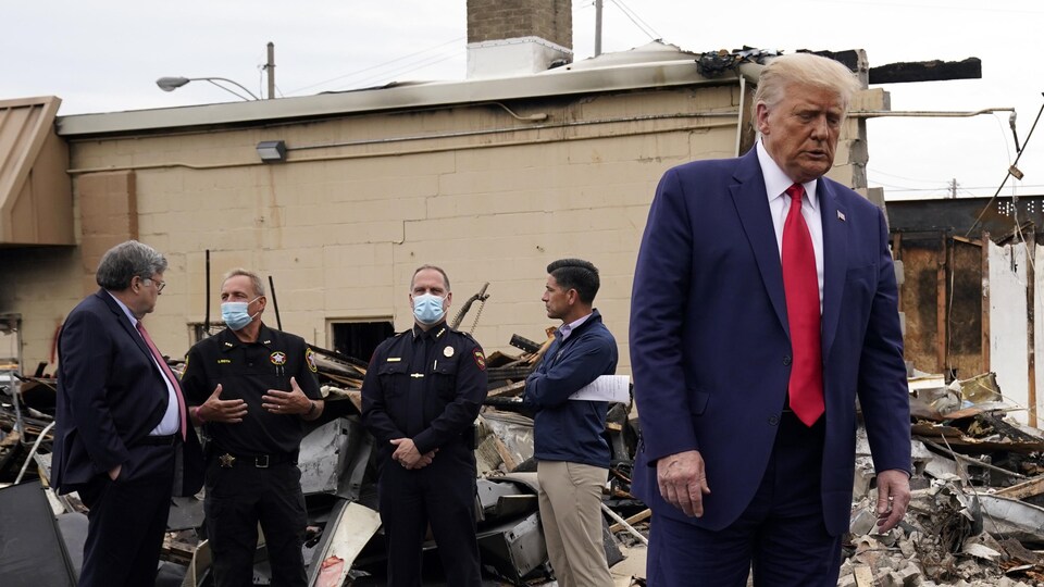 Donald Trump fait dos à des policiers près d'un immeuble détruit.