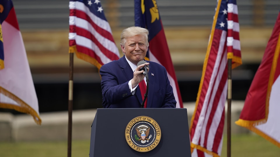 Donald Trump sourit en ajustant le micro debout à une tribune, devant des drapeaux des États-Unis.