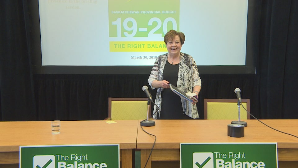 Donna Harpauer entre dans une salle de conférence, le sourire aux lèvres, avec un document dans ses mains. Elle est debout devant des micros. 
