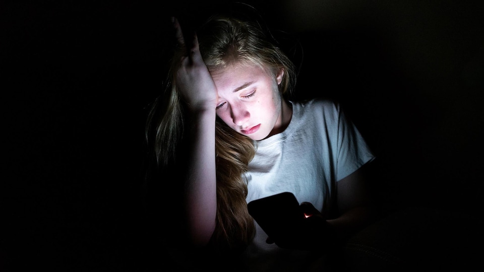 Une jeune fille regarde atterrée sont cellulaire dans le noir.