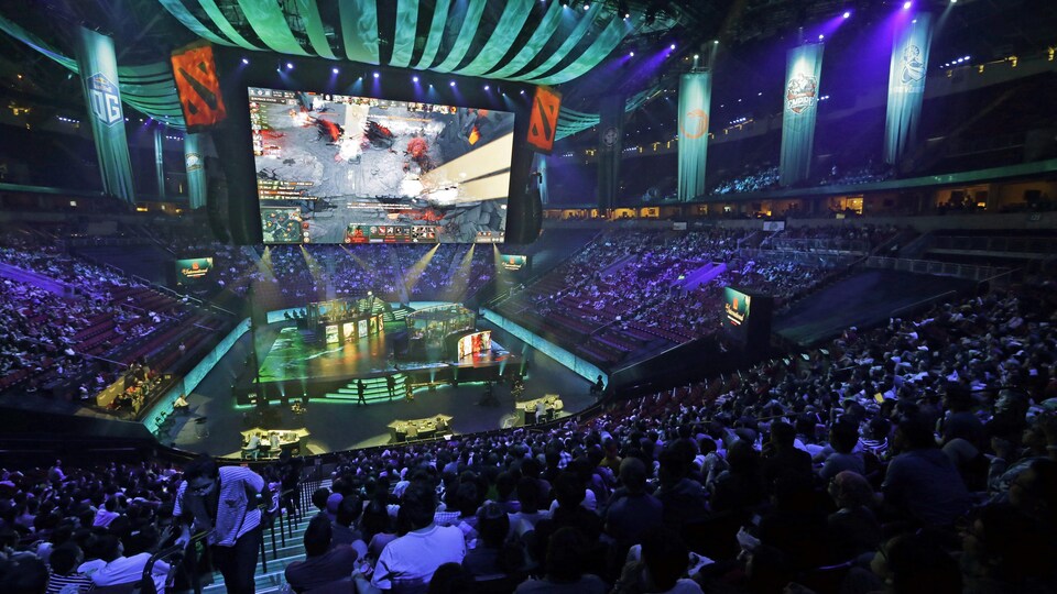 Une vue d'ensemble de l'intérieur d'un aréna, dans lequel des milliers de spectateurs sont assis dans les gradins, éclairés par une lumière violette tamisée. Au centre de l'aréna, d'immenses écrans géants sont suspendus devant une scène carrée et diffusent un match de Dota 2.