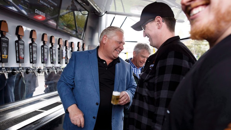 Le premier ministre Doug Ford, vêtu d'un blouson marine, avec un petit verre de bière à la main, des hommes souriants sont autour de lui et il se tient devant un comptoir de service de bière. 