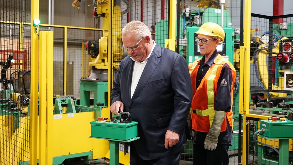 Le premier ministre Doug Ford examine une pièce d'équipement dans une usine en écoutant les explications d'une employée.
