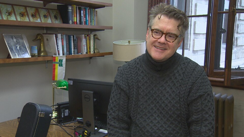 Dougald Lamont, assis sur son bureau, souriant. Au fond, on voit des étagères de livre et un drapeau franco-manitobain.