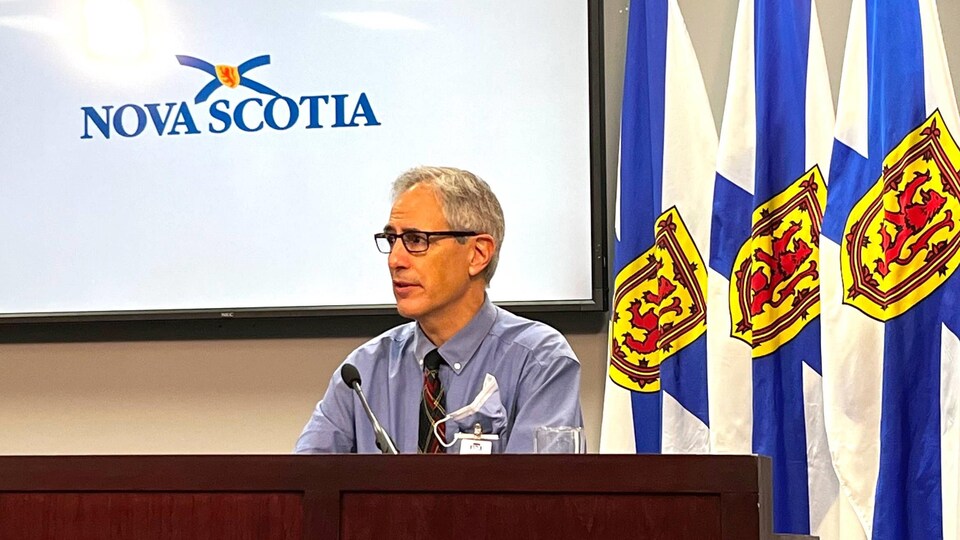 Un homme assis à une longue table durant une conférence de presse. Il y a un grand écran au mur derrière lui  avec le mot « Nova Scotia » qui apparaît, et des drapeaux de la Nouvelle-Écosse.