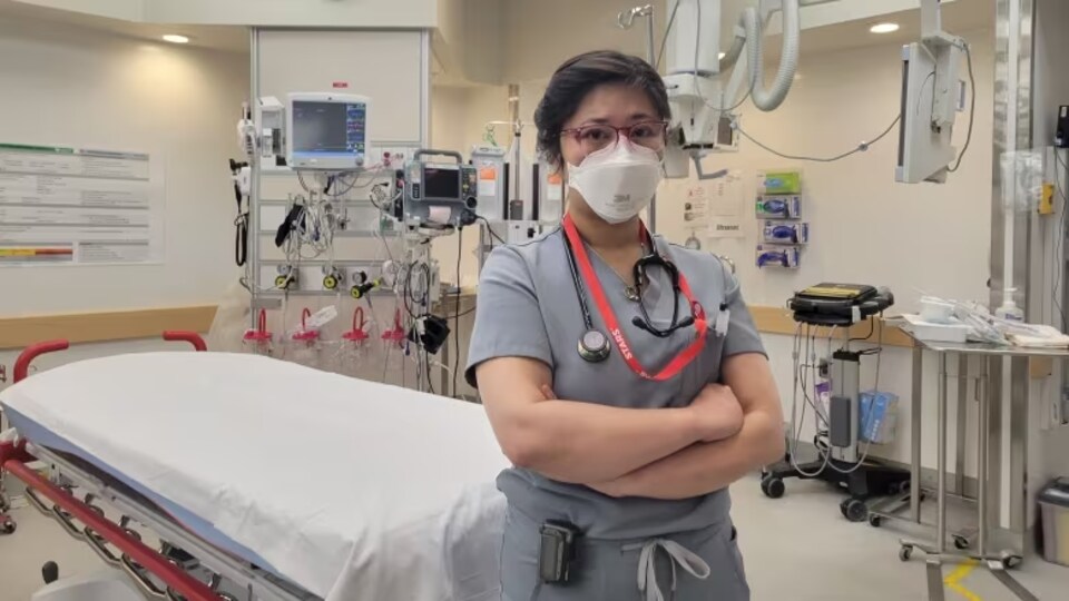Une médecin se tenant devant un lit d'hôpital, masque sur le visage et stéthoscope au cou.