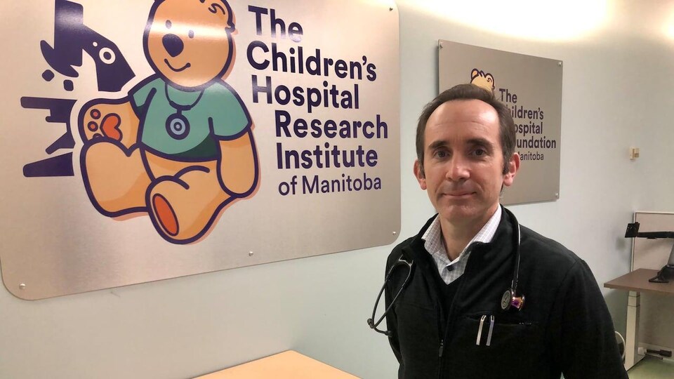 Le Dr Sergio Fanella est debout devant une affiche du centre de recherche sur les enfants, le Children's Hospital Research Institute of Manitoba.