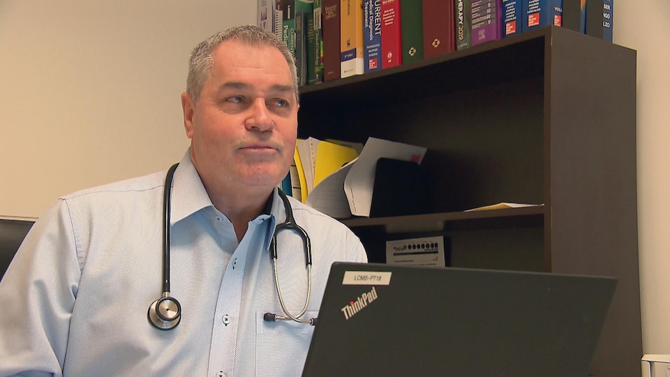 Dr. Bolduc was sitting at a desk, in front of a laptop computer, inside his office. He was wearing a stethoscope around his neck.