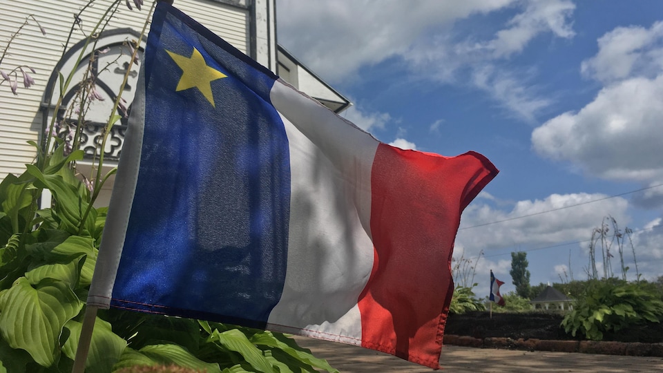 Un drapeau rouge, blanc et bleu avec une étoile jaune dans le coin gauche, planté dans une platebande de verdure devant un église blanche en bois.