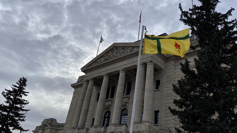 Le drapeau est hissé devant l'Assemblée législative de la Saskatchewan, à Regina. Le ciel est couvert avec quelques éclaircies en fond, et un sapin se tient à la droite du drapeau.