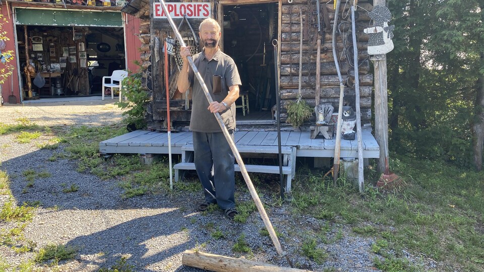 Un homme tenant une gaffe à la main pose devant une cabane en bois rond.