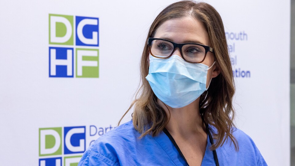 Une chirurgienne qui porte des lunettes pour la vue et son uniforme de travail a un masque chirurgical qui lui couvre le nez et la bouche. Elle est debout contre un panneau décoré du logo de l'Hôpital général de Dartmouth et elle regarde vers sa droite.