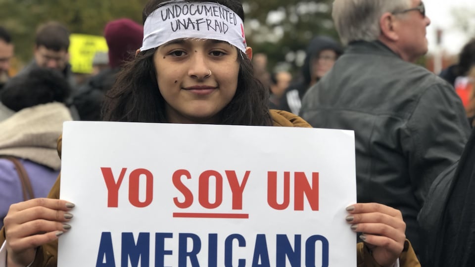 Gros plan d'une jeune femme qui tient dans ses mains une pancarte sur laquelle on peut lire : ''Yo soy un Americano'' (Je suis une Américaine).