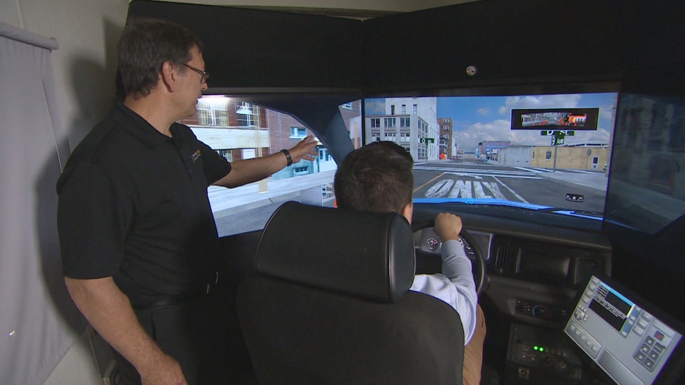 Un homme assis dans un simulateur de conduite, accompagné d'un instructeur qui pointe vers l'écran.