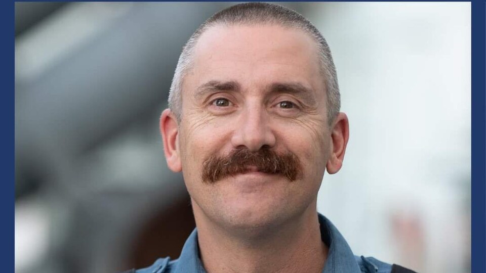 Duane Earle sourit. Il est vêtu de son équipement de travail formel, une chemise bleue.