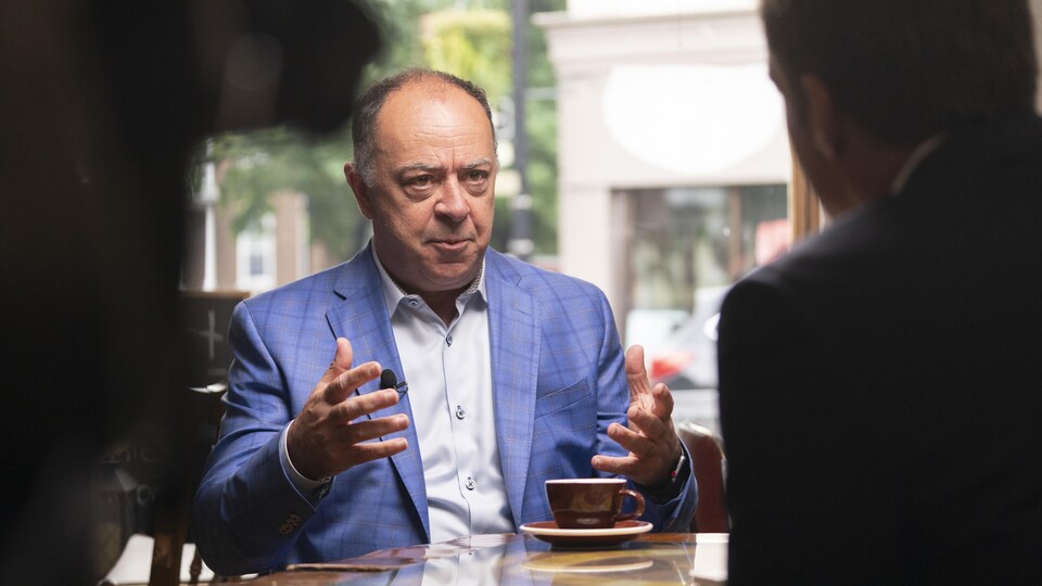 Le ministre Dubé, assis, de face, les mains ouvertes, en train de discuter avec l'animateur, qui est de dos. Ils sont assis autour d'un café. À gauche, on devine les équipements de télévision.
