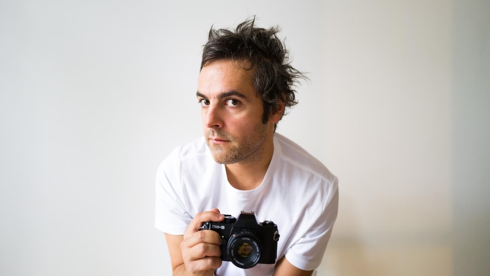 L'artiste pose devant un mur blanc avec un appareil photo dans la main. 