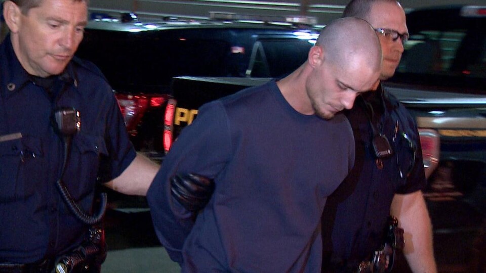 Un homme au crâne rasé et baissant la tête est tenu par deux policiers.