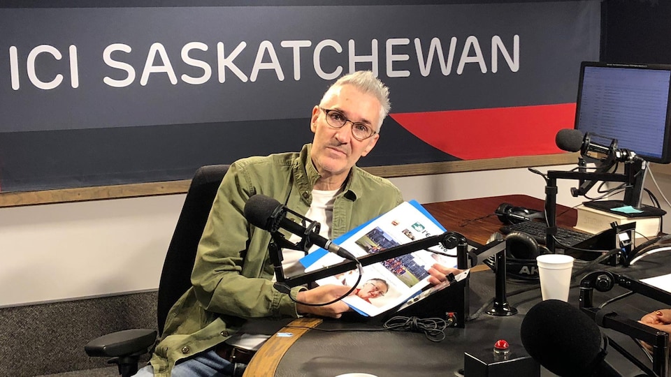 Un homme portant une veste verte et des lunettes présente la maquette de son journal dans le studio de radio de Radio-Canada.