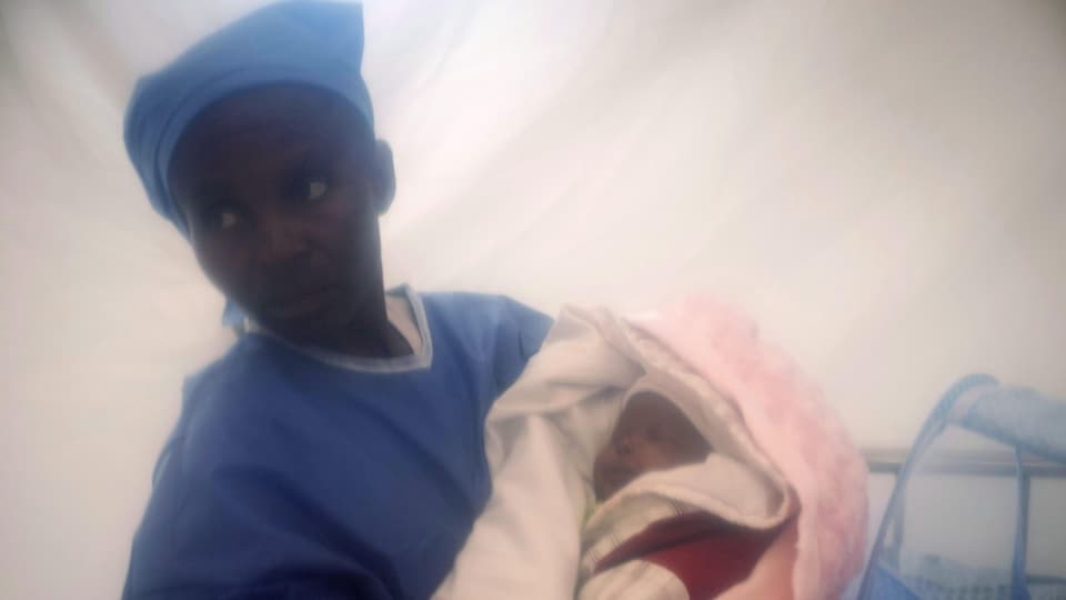 Une femme en jaquette d'hôpital tient un tout jeune enfant emmailloté, qui dort dans ses bras.