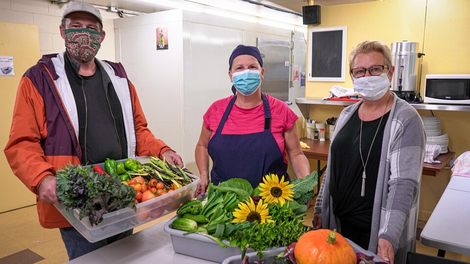 Trois personnes posent pour la caméra dans une cuisine. Ils tiennent dans leur mains des bacs remplis de légumes frais.