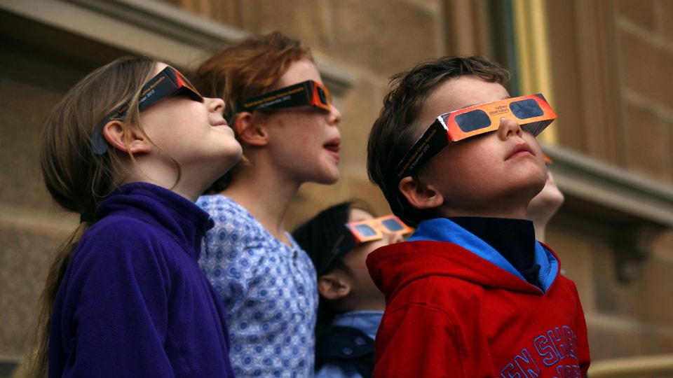 Quatre enfants avec des lunettes de protection orange et noire en carton. Leurs têtes sont levées vers les cieux.