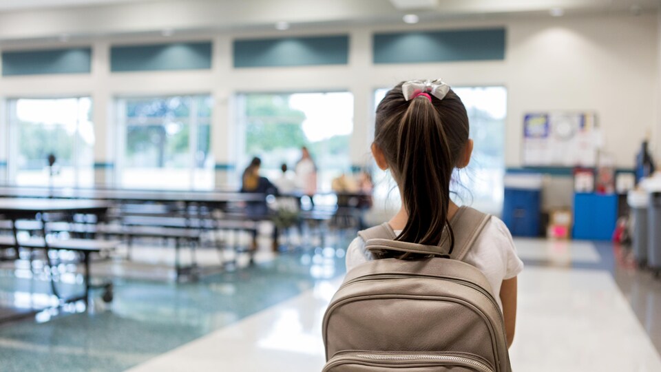 Une petite fille avec un sac à dos vue de dos alors qu'elle entre dans la cafétéria d'une école.